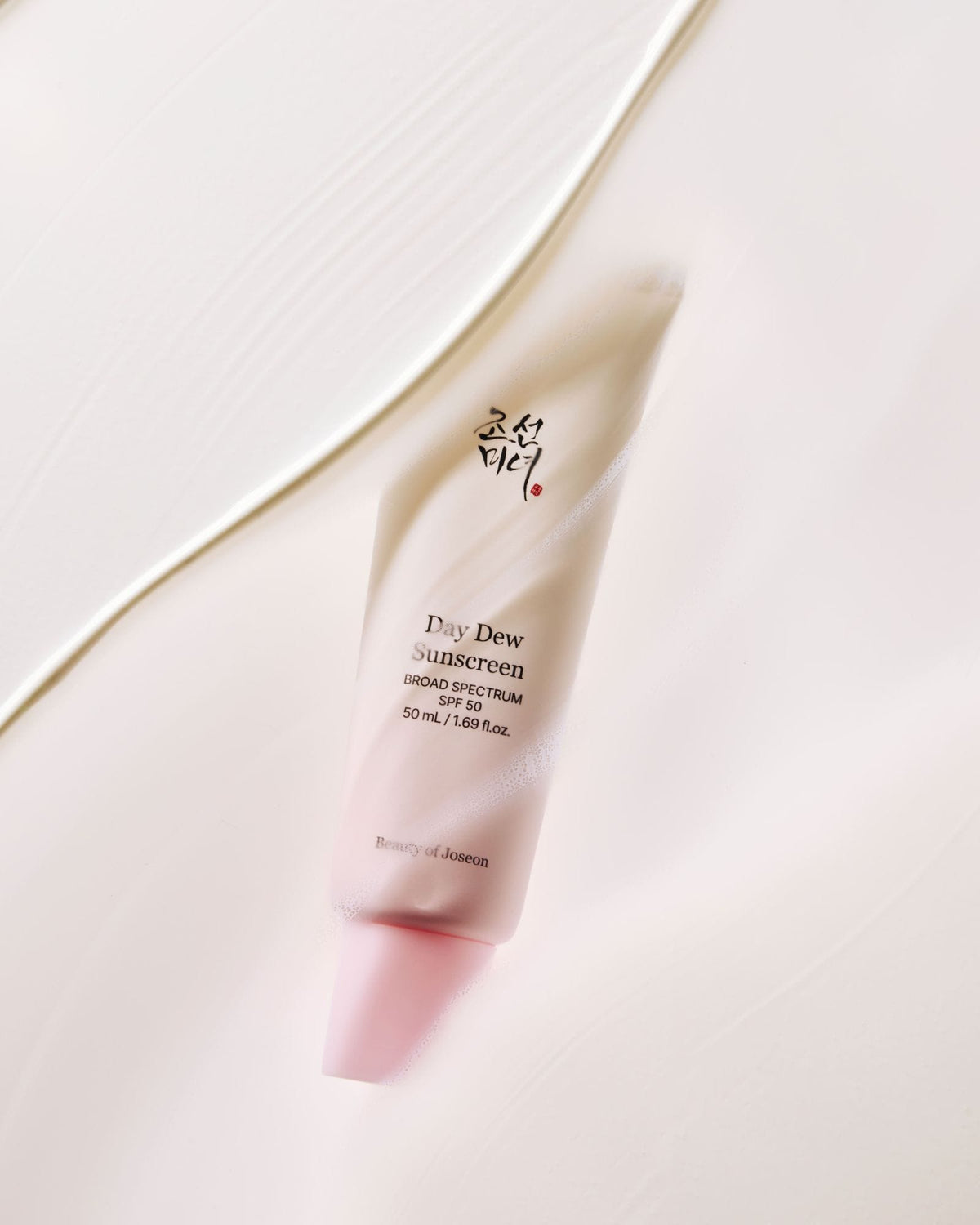 A squeeze tube of Day Dew Sunscreen by Beauty of Joseon, featuring a soft pink cap and a pale peach body with black text, set against a clean, light beige background with a diagonal gold line.