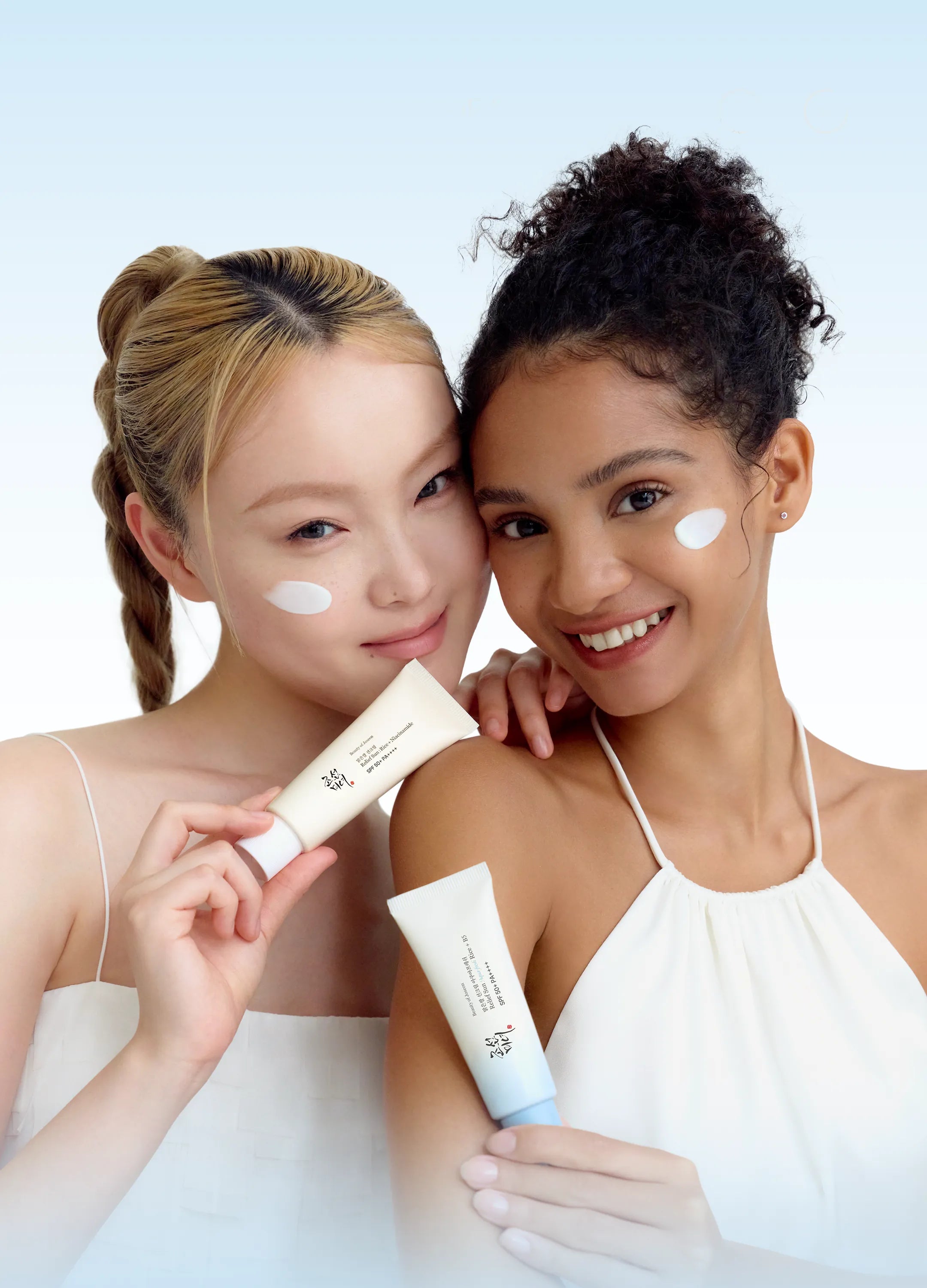 Two women smiling and holding sunscreen tubes, with white sunscreen cream visible on their cheeks, against a light blue gradient background.